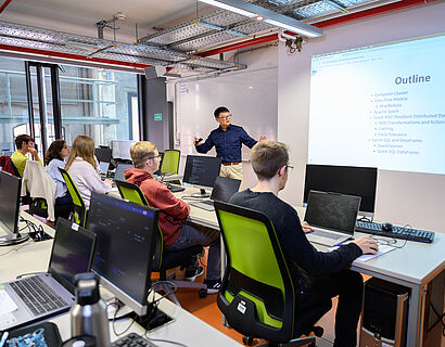 Blick in einen Seminarraum auf eine Sitzreihe mit Studierenden von hinten, vorne an der Tafel spricht und gestikuliert ein Professor. 