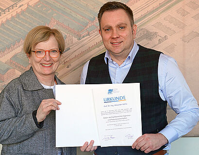 Frau und Mann stehen nebeneinander und halten eine Urkunde in der Hand.