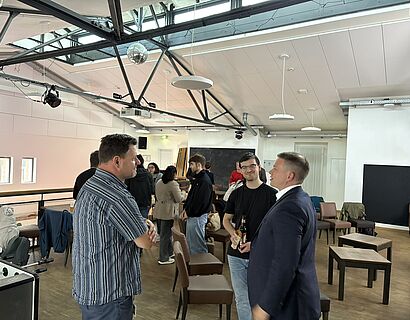 Blick in einen Raum, in dem sich Personen in losen Grüppchen unterhalten. Im Vordergrund sprechen drei Personen, einer hat eine Getränk und ein Gebäckstück in der Hand.