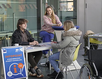 An einer Tischgruppe sitzen drei Personen und unterhalten sich. Ein Aufsteller im Vordergrund weist auf den "Infopoint" hin, ebenso ist ein Rollstuhl zu sehen.