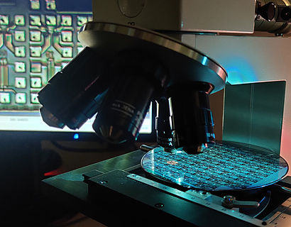 Mikrochips auf einem Halbleiter-Wafer unter dem Lichtmikroskop. Im Hintergrund zeigt ein Monitor die vergrößerte Ansicht.
