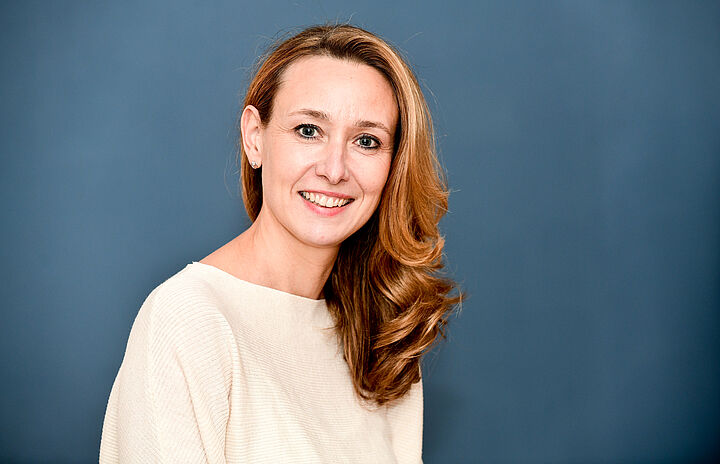 Porträtaufnahme von Marina Dillmann lächelnd vor einem blauen Hintergrund.