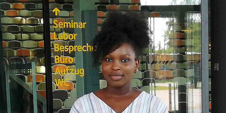 Junge Frau steht vor einem Hörsaal mit farbigen Sitzen und schaut in die Kamera