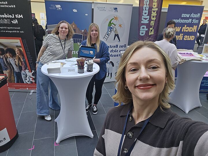 Selfie einer Mitarbeiterin des Karrierezentrums für professorale Entwicklung auf einer Karrieremesse. Im Hintergrund sind zwei weitere Mitarbeiterinnen des Karrierezentrums an einem Messestand zu sehen.