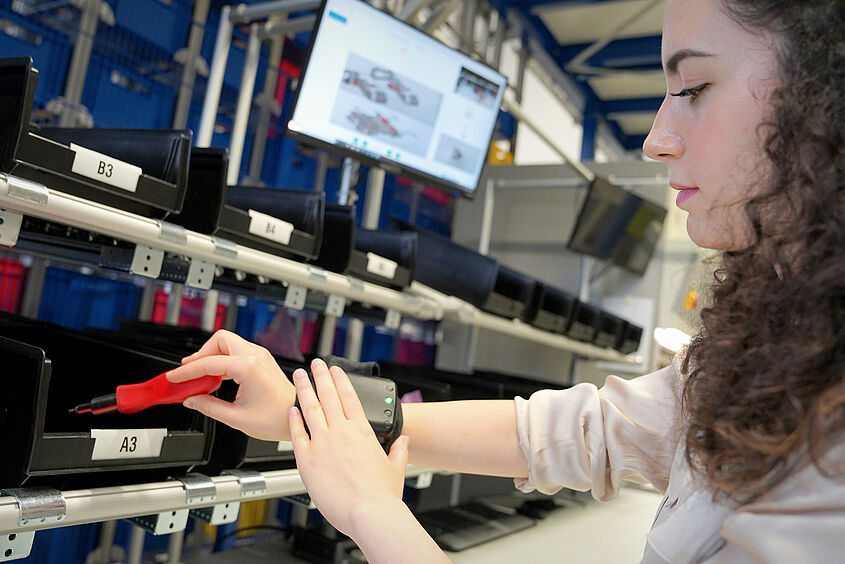 Studierende steht im Intralogistiklabor am Warenregal und synchronisiert den Bestand mit einem Sender am Handgelenk