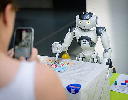 Humanoider Roboter beim Autogramm-Schreiben wird von einer Besucherin mit dem Smartphone fotografiert.