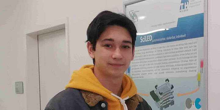 Junger Mann mit schwarzen kurzen Haaren steht vor einem Poster an der Wand und schaut in die Kamera