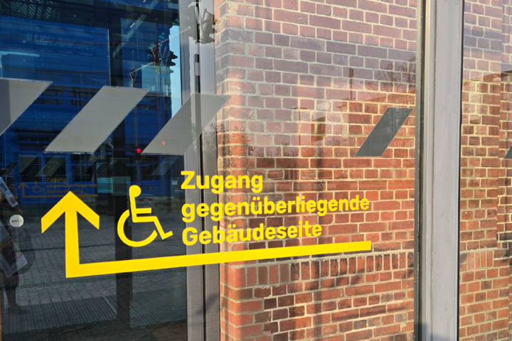 Man sieht ein barrierefreies Schild für einen Zugang von Halle 14 auf der gegenüberliegenden Seite