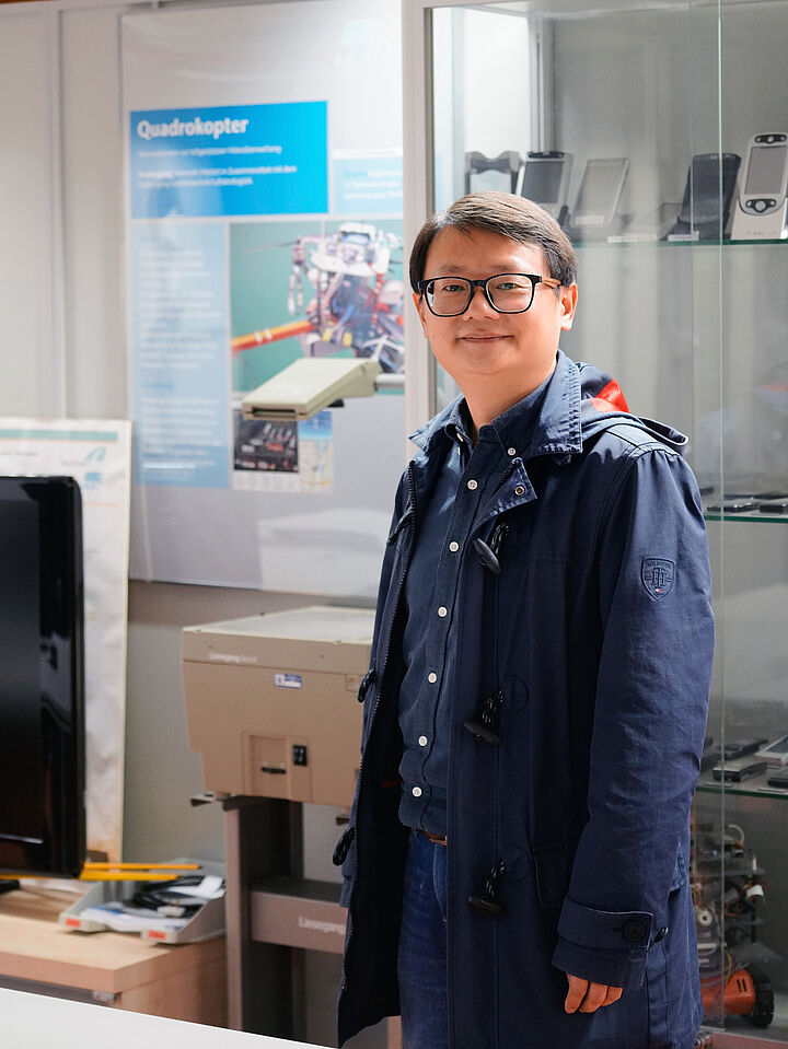 Ein lächelnder Professor steht in einem Labor für Telematik vor einer Glasvitrine mit Mobiltelefonen.