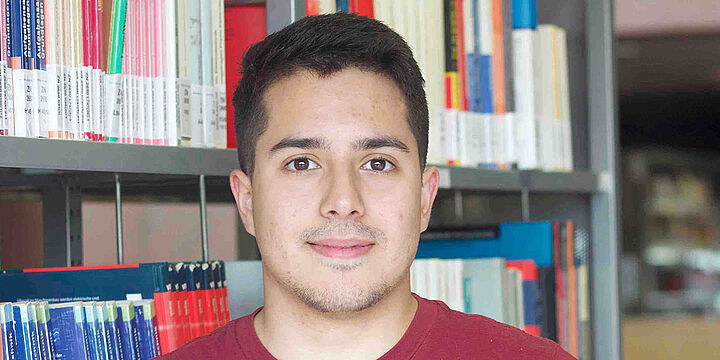 Junger Mann mit rotem Shirt steht vor einem Bücherregal