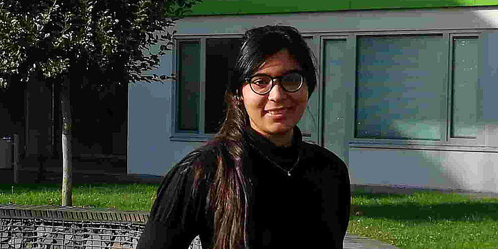 Junge Frau mit dunklen Haaren und schwarzem Pulli steht auf einer grünen Wiese vor einem Gebäude