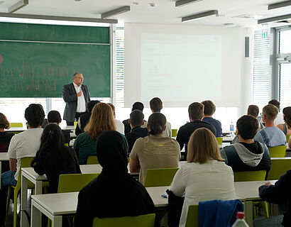 Von hinten fotografierte Studierende hören in einem Seminarraum den Ausführungen eines Dozierenden zu