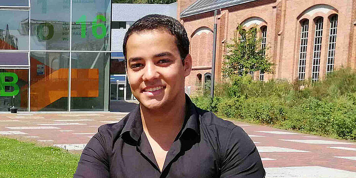 Junger Mann sitzt auf einer Bank auf dem Campus mit roten Backsteingebäuden im Hintergrund