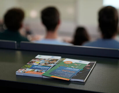 Informationsbroschüren liegen auf einem Tisch im Hörsaal mit Studierenden im Hintergrund