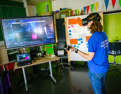 Besucher des HIT 2024 hat eine VR-Brille auf dem Kopf und steht vor einem mobilen Bildschirm.