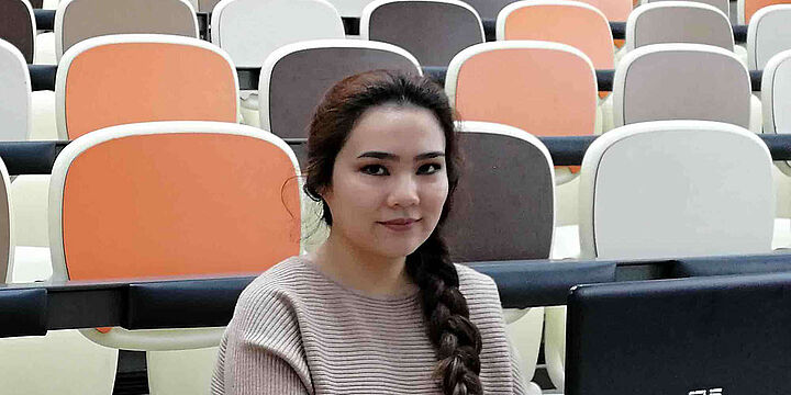 Junge Frau mit dunklen langen Haaren sitzt in einem Horsaal mit unterschiedlich farbigen Stühlen