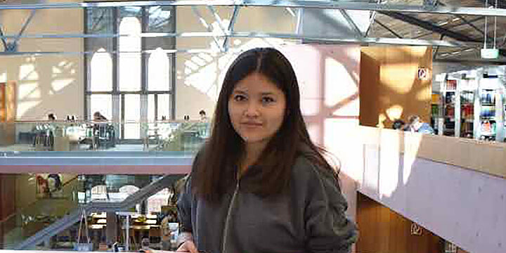 Junge Frau steht in einer oberen Etage der Hochschulbibliothek und schaut in die Kamera