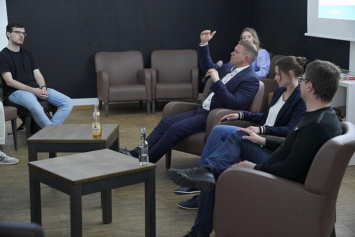 Blick von der Seite auf ein Podium mit drei Speaker*innen, ein Redner spricht ins Mikrofon und gestikuliert.