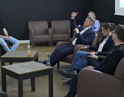 Blick von der Seite auf ein Podium mit drei Speaker*innen, ein Redner spricht ins Mikrofon und gestikuliert.