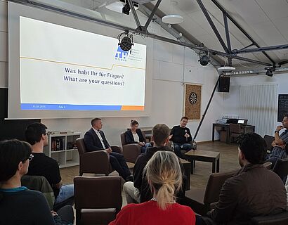 Blick von hinten auf zwei Publikumsreihen, die nach vorne auf ein Podium mit drei Personen schauen.