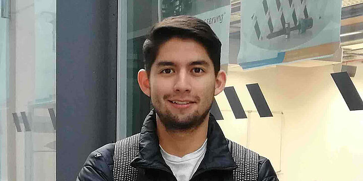 Junger Mann steht vor einer Glasscheibe hinter der sich ein Seminarraum in einem Hörsaalgebäude befindet