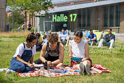 Zwei junge Frauen und ein junger Mann sitzen auf einer Wiese und schauen gemeinsam auf ein Tablet