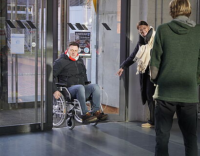 Ein Mann rollt im Rollstuhl durch eine Tür, während eine Frau auf die Tür deutet.