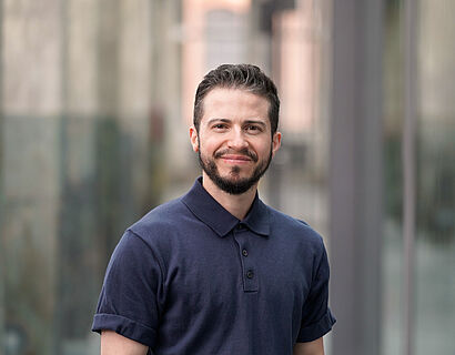 Person mit dunklem Shirt, schwarzen Haaren und Bart steht für ein Portraitbild in einer alten Industriehalle mit Betonpfeilern im Hintergrund.